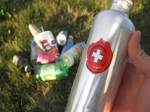 A reusable water bottle in a hand, on the ground behind the bottle are a pile of discarded single use plastic water bottles