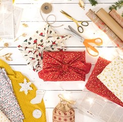 A picture of two having been wrapped in fabric and surrounded by rolls of wrapping paper and a selection of fabric, scissors and string.